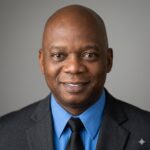 Smiling African American man in a dark suit, blue shirt, and black tie, posing for a portrait against a gray background.