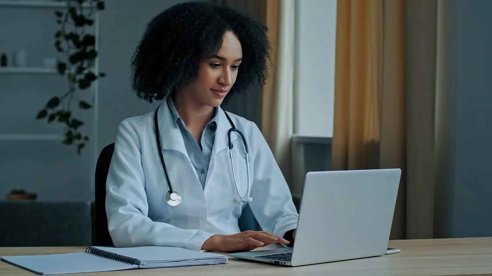 Female doctor in a white coat with a stethoscope sits at a desk, typing on a laptop with a notebook nearby.